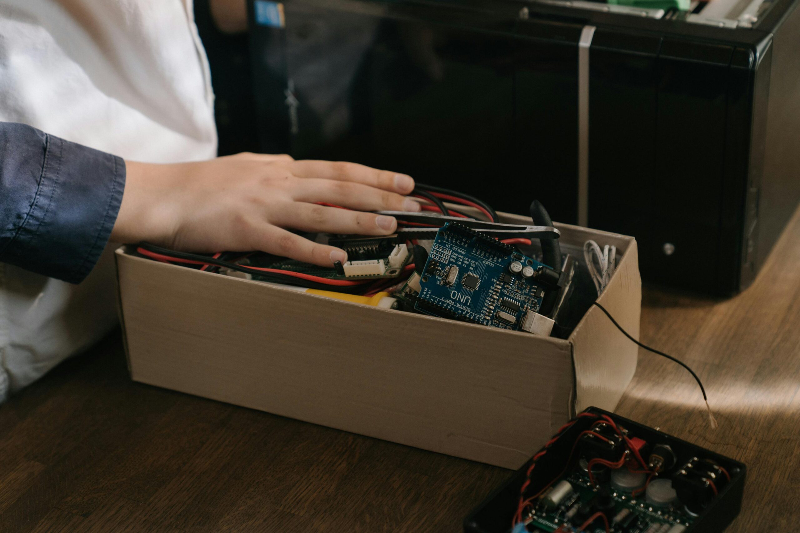 Osoba trzyma rękę na pudełku z elementami elektronicznymi, w tym płytką Arduino UNO i przewodami.